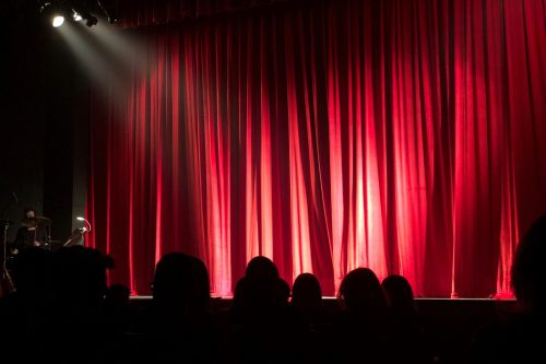 Dimly lit theater stage with red curtains and audience silhouettes under spotlights.